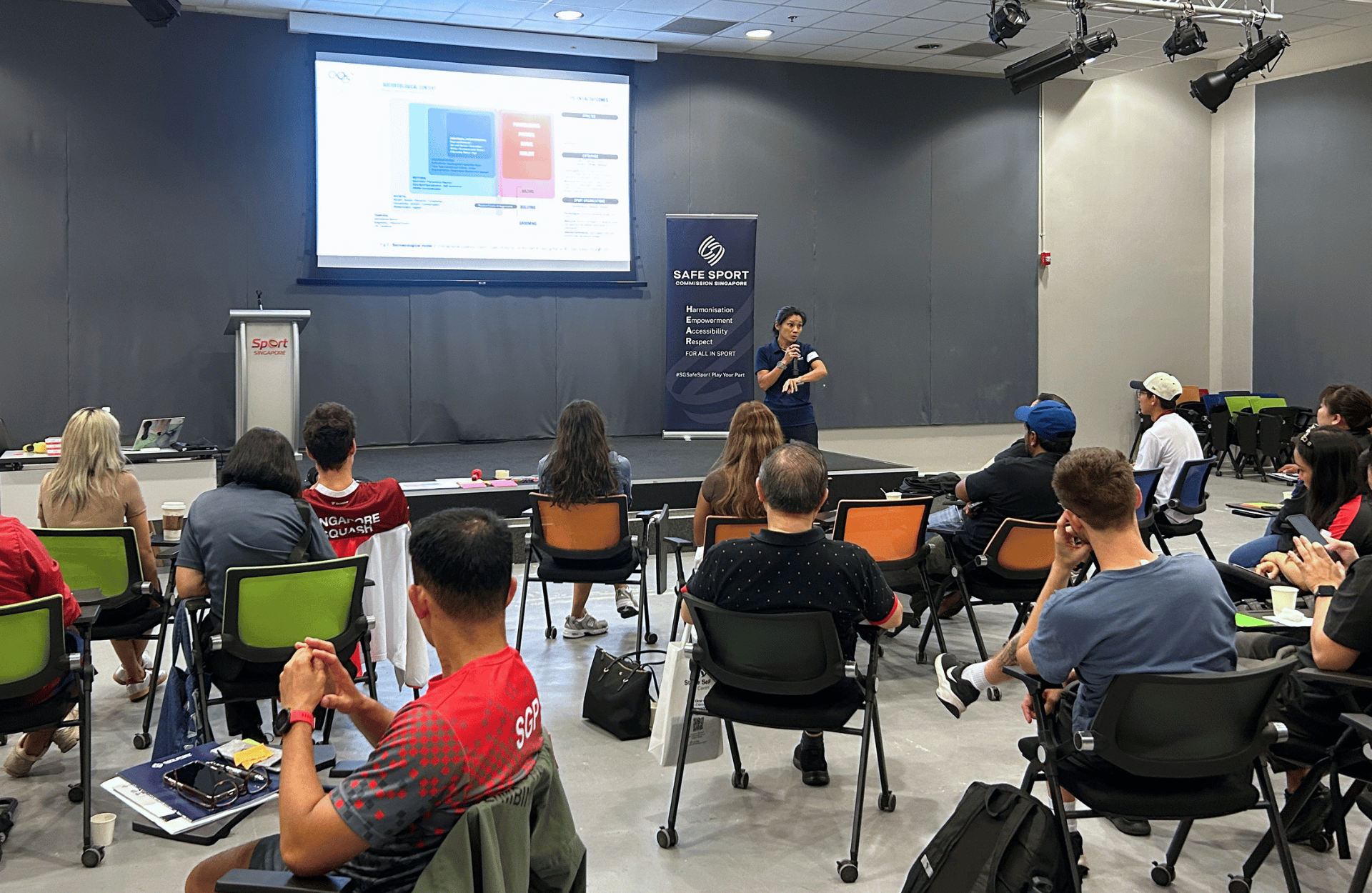 A speaker addresses a group of attentive listeners during a presentation, with visual aids displayed behind her.